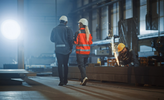 Two people in hard hats and safety clothing walking past a welder in a manufacturing setting. 