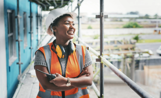 Worker in a hard hat and orange safety vest holding a walkie talkie and smiling as she looks into the distance, standing on a scaffold. 