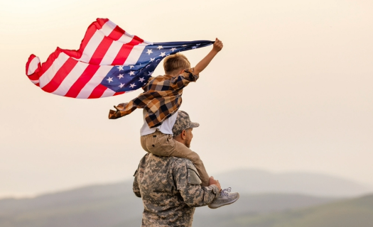 A soldier with a child riding on his shoulders waving an American flag.
