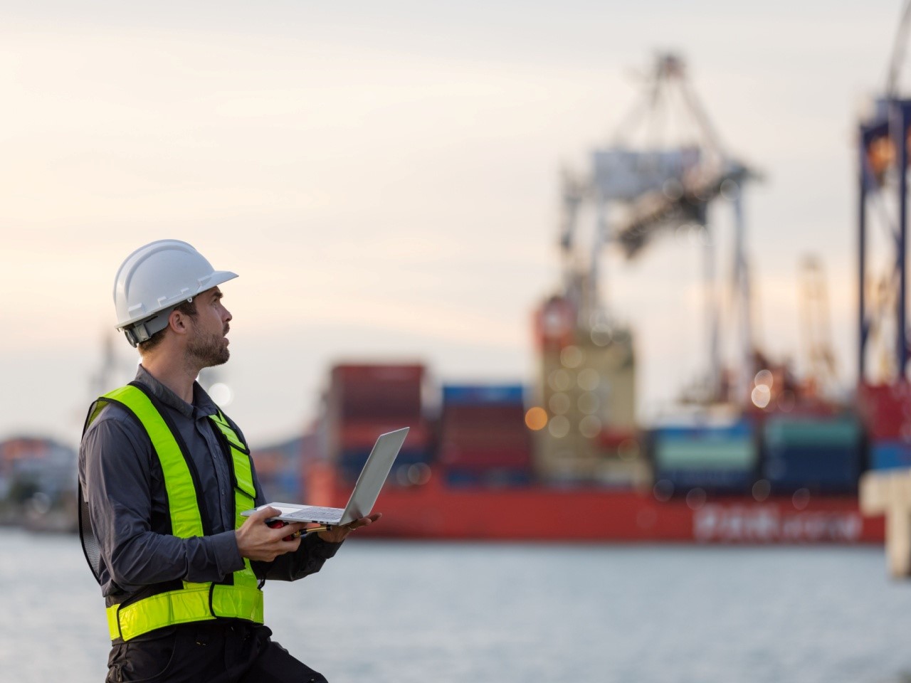 A male engineer in a safety vest and helmet uses a laptop at a busy shipping port, with cargo containers and cranes in the background, symbolizing logistics, transportation, and maritime operations.