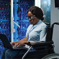 African female IT engineer with disability sitting in wheelchair and typing data on laptop during her work in data center